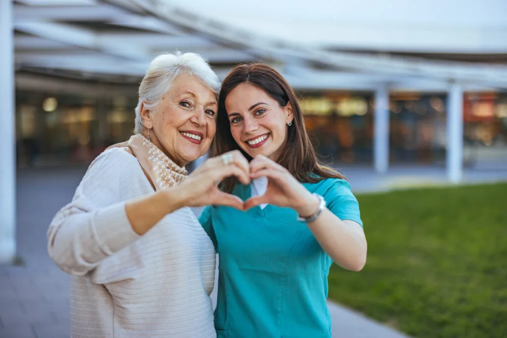 An older woman and her caregiver making a heart shape with their hands